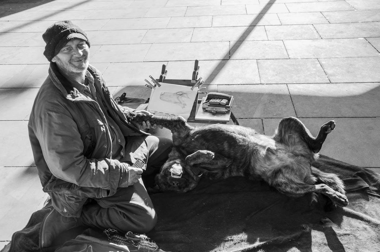 A joyful street artist and his dog relax on a London pavement as he sketches.
