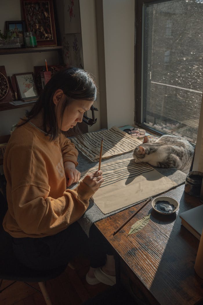 Artistic shot of an adult woman practicing calligraphy near a resting cat by a sunlit window in New York.